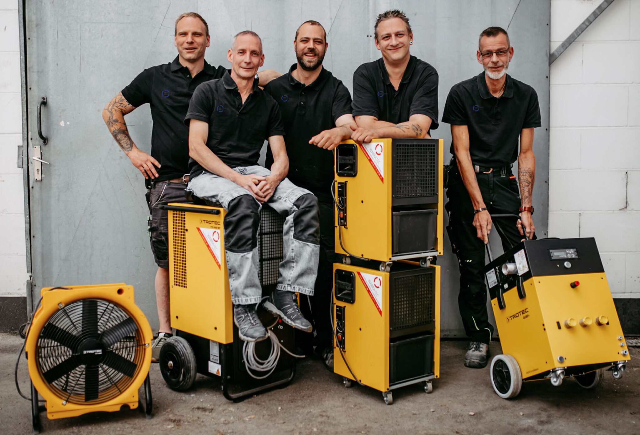Fünf Handwerker in schwarz weiß posieren mit gelben Geräten für die professionelle Bautrocknung.